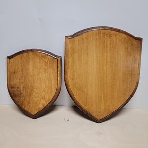 May include: Two wooden shield plaques with a light brown finish and dark brown trim. The plaques are in a shield shape, with a curved top and pointed bottom. The wood grain is visible.