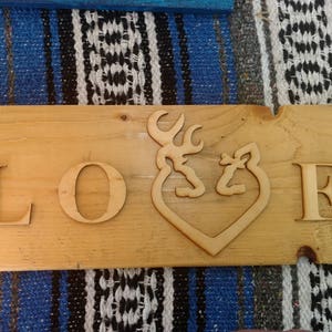 May include: Wooden sign with the word "LOVE" carved into it. The "O" is replaced with a heart-shaped design featuring a deer and a buck. The sign is made of light-colored wood.