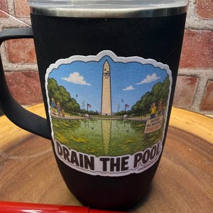May include: A black insulated mug with a handle and a clear lid, featuring a sticker of the Washington Monument with a sad face, a green reflecting pool, and the text "DRAIN THE POOL" and "ANOTHER BIG BEAUTIFUL DISASTER" printed on the front.