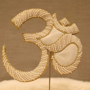 May include: A gold-coloured Om symbol made of white beads and a white fabric backing. The symbol is attached to a gold-coloured spring stand.