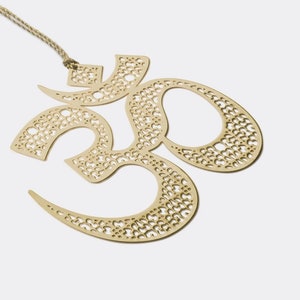 May include: A gold-colored metal Om symbol ornament with a delicate, intricate design. The symbol is suspended from a length of twine.