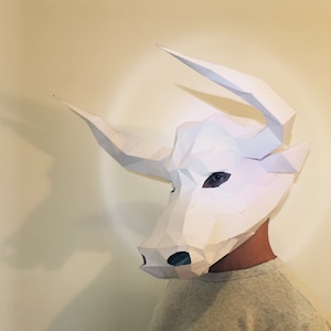 Könnte beinhalten: Eine weiße Papier-Stiermaske mit geometrischem Design. Die Maske wird von einer Person getragen, aber das Gesicht der Person ist nicht sichtbar.