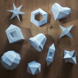 May include: A collection of white geometric paper ornaments in various shapes, including stars, a heart, a hexagon, a bell, and a sphere. The ornaments are arranged on a wooden surface, creating a modern, minimalist aesthetic.