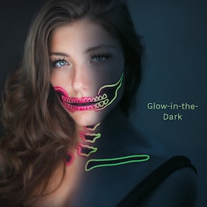 以下が含まれることがあります： 長い茶色の髪の女性が、暗闇で光るメイクをしています。メイクは頭蓋骨の形をしていて、緑とピンクに光っています。テキスト「Glow-in-the-Dark」は、青い背景に白で書かれています。
