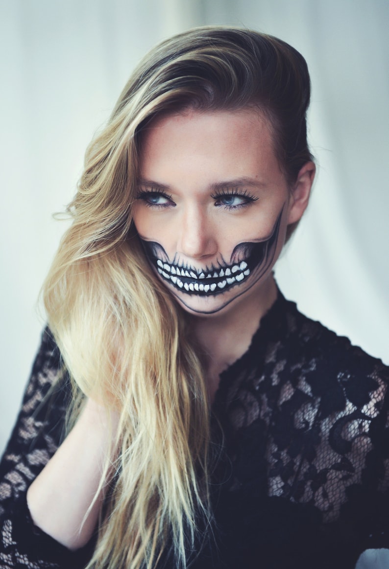 May include: A woman with long blonde hair wearing a black lace top has a skull painted on her face. The skull is black and white with a white smile.