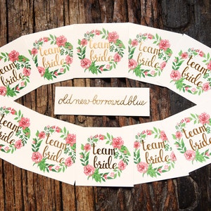 Op de afbeelding: Negen witte vierkante kaarten met gouden letters die "team bride" zeggen in een bloemkrans. De kaarten liggen op een houten oppervlak. De tekst "old new borrowed blue" staat op een aparte kaart.