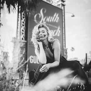 May include: A woman with curly hair and a skeleton face paint sits in front of a sign that reads "South Beach".