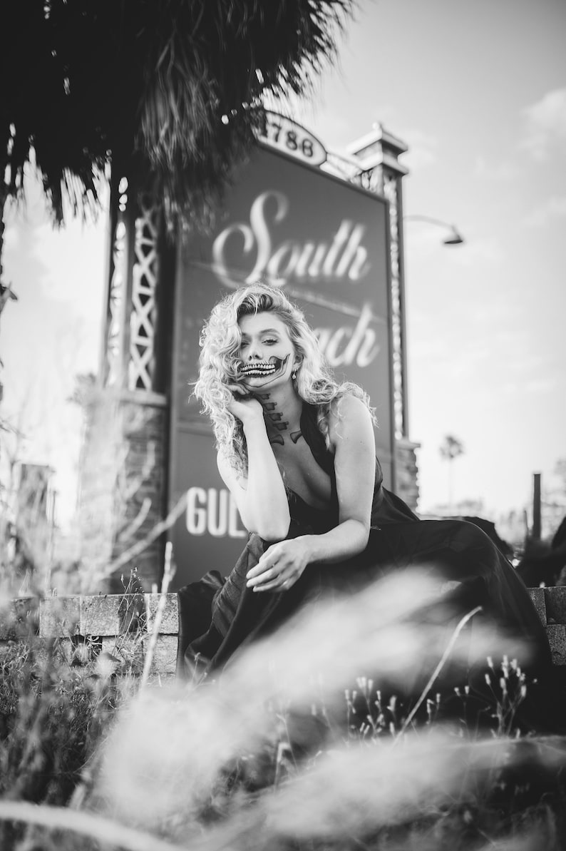 May include: A woman with curly hair and a skeleton face paint sits in front of a sign that reads "South Beach".