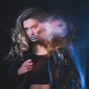 May include: A woman with blonde hair wearing a black leather jacket and a skull makeup design on her face. The image is blurred and has a blue and white light effect.