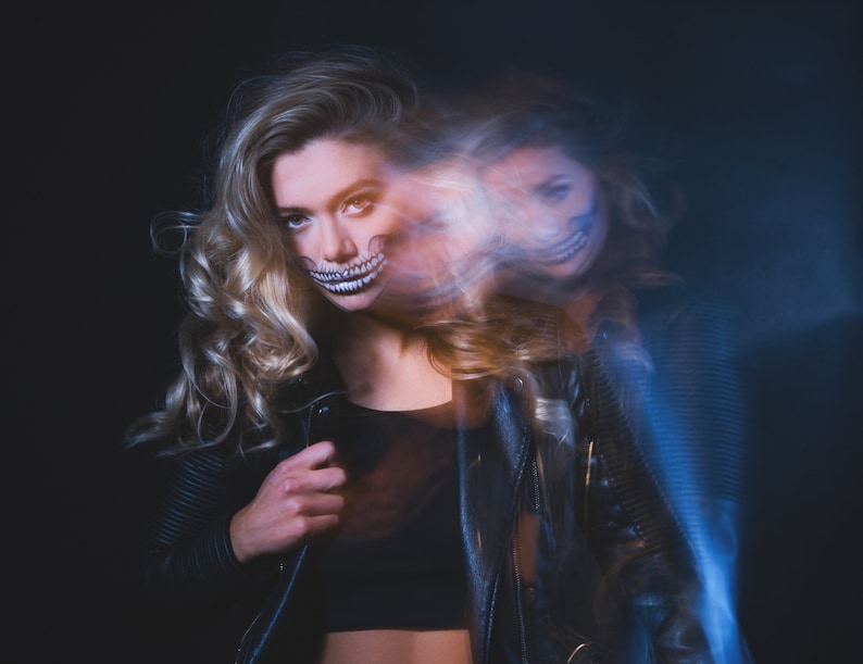 May include: A woman with blonde hair wearing a black leather jacket and a skull makeup design on her face. The image is blurred and has a blue and white light effect.