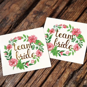 Könnte beinhalten: Zwei weiße quadratische temporäre Tattoos mit einem floralen Kranzdesign und goldener Aufschrift "team bride".