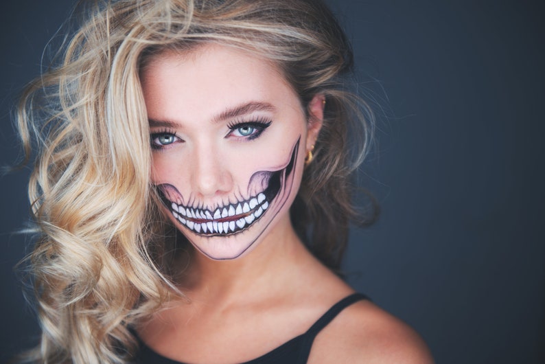 May include: A woman with blonde hair and a skull face paint. The paint is black and white and covers her entire face. She is wearing a black tank top.