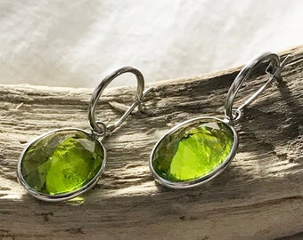 May include: A pair of silver hoop earrings with green oval-shaped gemstones hanging from them. The earrings are displayed on a piece of driftwood.