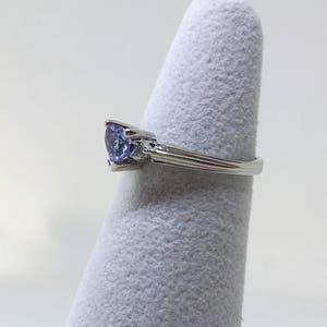 K&ouml;nnte beinhalten: Ein silberner Ring mit einem herzf&ouml;rmigen blauen Edelstein und kleineren klaren Edelsteinen auf der Ringschiene.