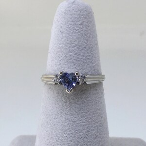 K&ouml;nnte beinhalten: Ein silberner Ring mit einem herzf&ouml;rmigen blauen Edelstein und zwei kleineren klaren Edelsteinen auf der Ringschiene.