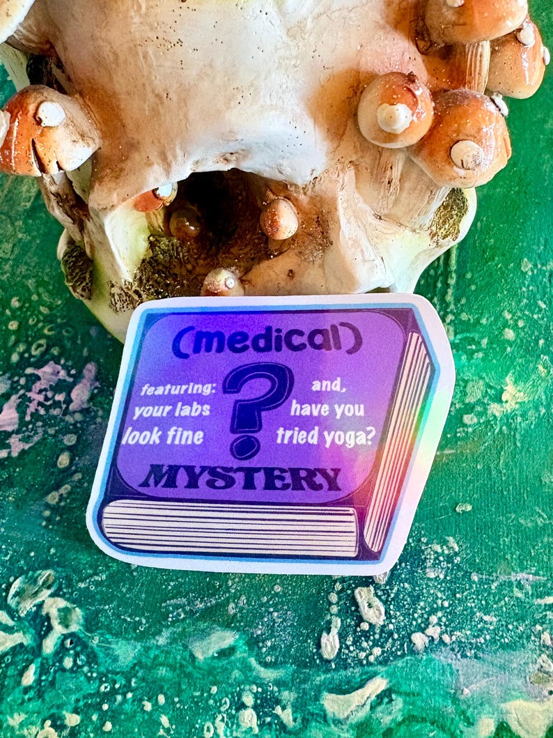 May include: A holographic sticker with the text "(medical)", "featuring: your labs look fine", a question mark, and "and, have you tried yoga?" The word "MYSTERY" is also on the sticker. The sticker is on a green and white surface.
