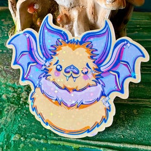 May include: A sticker featuring a cartoon bat with large, detailed wings. The bat has a light yellow body, purple accents, and blue wings with a holographic sheen. The background includes a skull and mushroom decor.