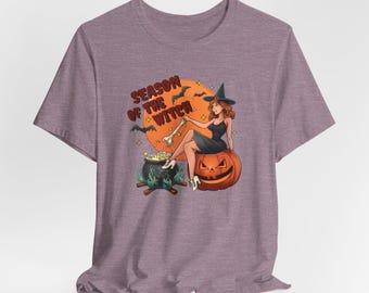 May include: Purple t-shirt with a graphic of a witch stirring a cauldron with a bone. The witch is sitting on a pumpkin and the text "Season of the Witch" is above her.