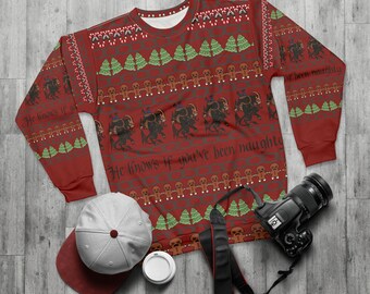 May include: Red Christmas sweater with a repeating pattern of gingerbread men, Christmas trees, and Krampus figures. The text "He knows if you've been naughty" is printed on the front. A gray baseball cap and a camera are next to the sweater.