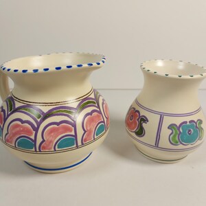 May include: Two cream-coloured ceramic vases with hand-painted floral designs. One vase has a handle and a rounded shape, the other is smaller and cylindrical. Both have blue, pink, and green accents.