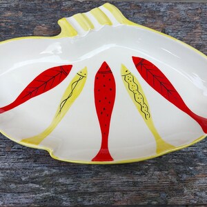 May include: A white ceramic serving dish shaped like a seashell with a yellow rim. The dish is decorated with five red and yellow fish.