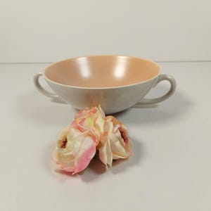 May include: A light grey ceramic soup bowl with two handles and a light peach interior. The bowl is sitting on a white surface with three light pink and white flowers.