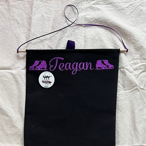 May include: Black fabric hanging organizer with the name "Teagan" embroidered in purple glitter. The organizer has two purple glitter ice skate images on either side of the name. A small white button with a skating image and the text "Championships 2023" is attached to the organizer.