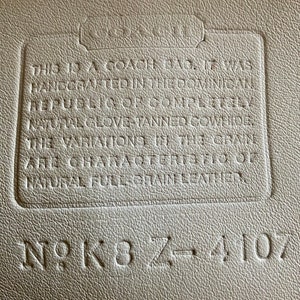 May include: Close-up of a white leather tag with the text "This is a Coach bag. It was handcrafted in the Dominican Republic & of completely natural glove-tanned cowhide. The variations in the grain are characteristics of natural full-grain leather." and the serial number "N9K8Z-4107".