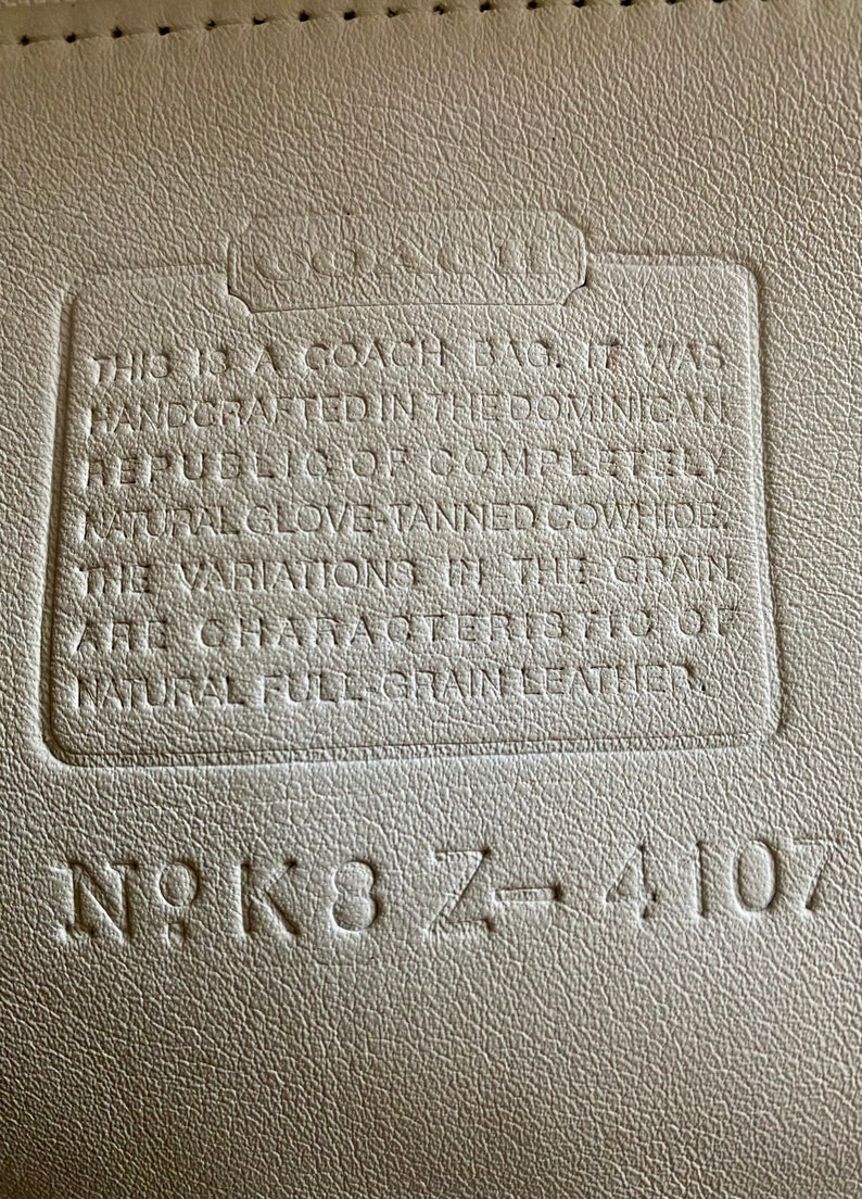 May include: Close-up of a white leather tag with the text "This is a Coach bag. It was handcrafted in the Dominican Republic & of completely natural glove-tanned cowhide. The variations in the grain are characteristics of natural full-grain leather." and the serial number "N9K8Z-4107".