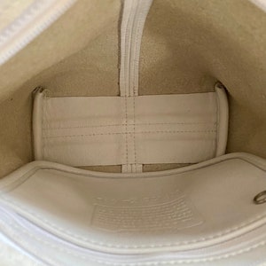 May include: The inside of a white leather bag with a white fabric lining. The bag has a zipper closure and a small pocket on the inside. The bag is empty.