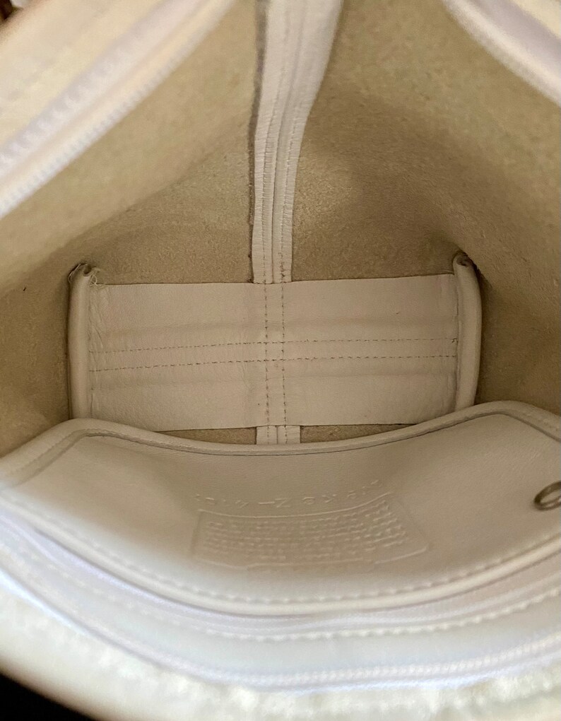 May include: The inside of a white leather bag with a white fabric lining. The bag has a zipper closure and a small pocket on the inside. The bag is empty.