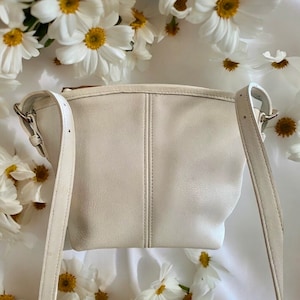 May include: A white leather crossbody bag with a single strap. The bag has a simple, minimalist design and is made of a soft, supple leather.