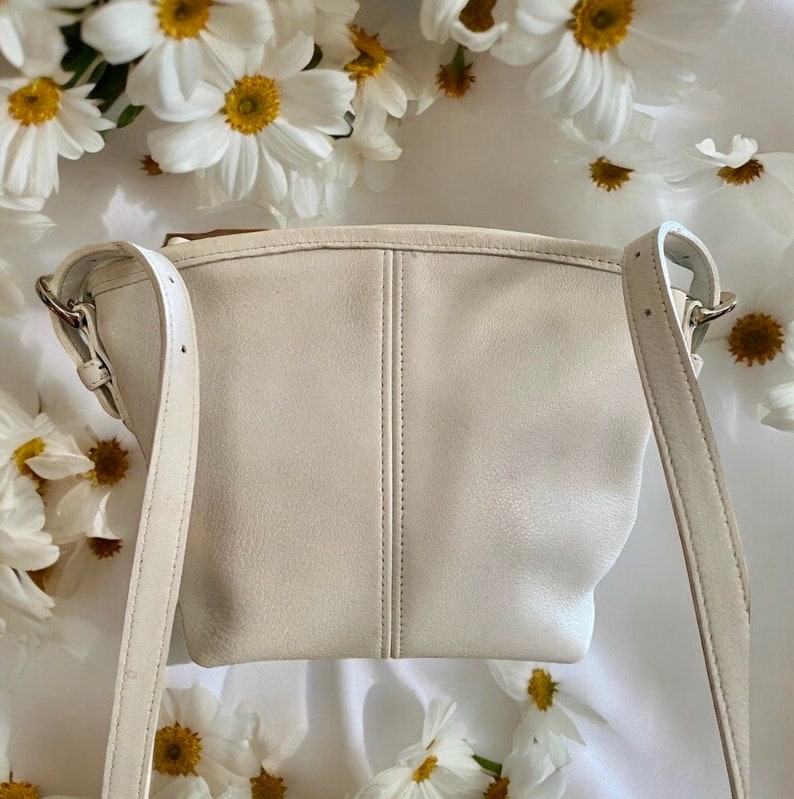 May include: A white leather crossbody bag with a single strap. The bag has a simple, minimalist design and is made of a soft, supple leather.
