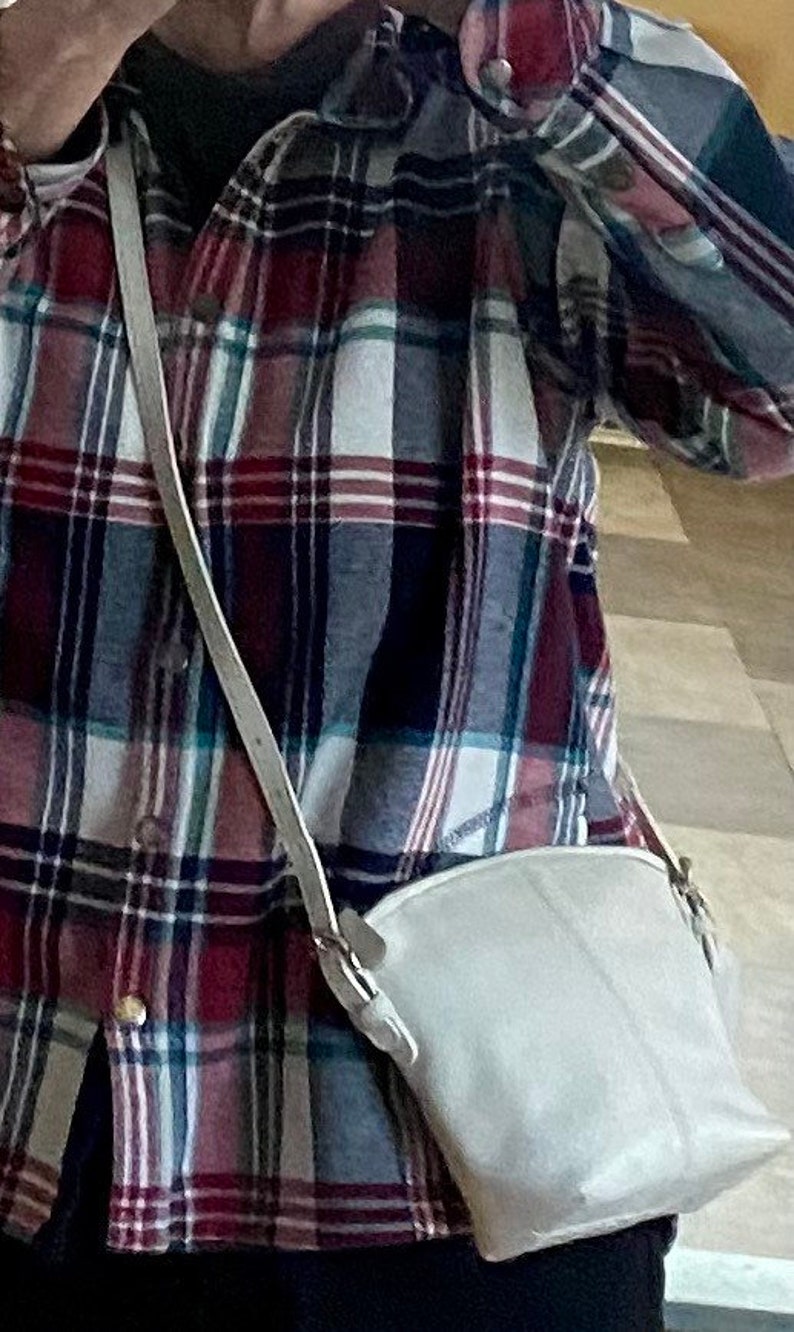 May include: A white crossbody bag with a long strap worn over a red, white, and blue plaid shirt.