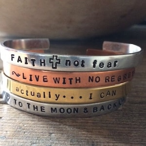 May include: Four stacked silver, copper, and gold bracelets with inspirational messages engraved on them. The top bracelet reads "FAITH not fear", the second bracelet reads "LIVE WITH NO REGRET", the third bracelet reads "actually... I CAN", and the bottom bracelet reads "TO THE MOON & BACK".
