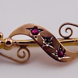 Op de afbeelding: Een goudkleurige broche met een sierlijk, vloeiend ontwerp. De broche heeft een centraal ribbonelement met drie rode edelstenen en steraccenten. Het ontwerp bevat spiraal- en bolvormige details.