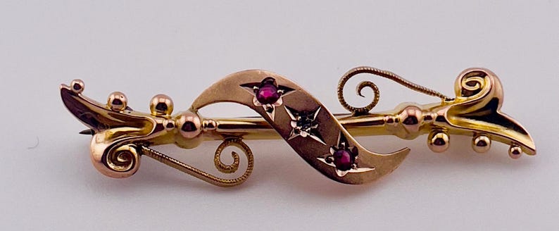 Op de afbeelding: Een goudkleurige broche met een sierlijk, vloeiend ontwerp. De broche heeft een centraal ribbonelement met drie rode edelstenen en steraccenten. Het ontwerp bevat spiraal- en bolvormige details.