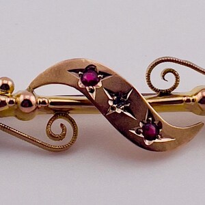 Op de afbeelding: Goudkleurige broche met een sierlijk ontwerp, met drie rode edelstenen en steraccenten. De broche heeft een speldsluiting en is waarschijnlijk vintage of antiek.