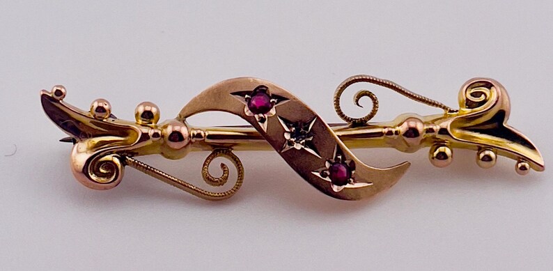 Op de afbeelding: Goudkleurige broche met een sierlijk ontwerp, met drie rode edelstenen en steraccenten. De broche heeft een speldsluiting en is waarschijnlijk vintage of antiek.