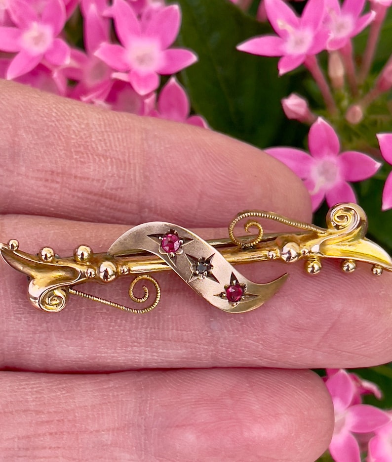 Op de afbeelding: Goudkleurige broche met een gebogen ontwerp, met een centraal lint versierd met rode edelstenen en een donkere edelsteen. De broche heeft ingewikkelde details en een decoratieve, scroll-achtige vorm. De achtergrond bevat roze bloemen.