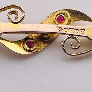 Op de afbeelding: Een vintage goudkleurige broche met een gebogen, vloeiend ontwerp. De broche heeft twee rode edelstenen en delicate krullen. De achterkant van de broche heeft een speldsluiting en gestempelde markeringen.