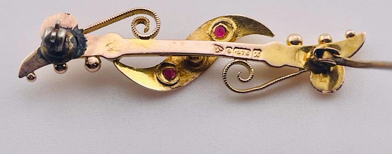 Op de afbeelding: Een vintage goudkleurige broche met een gebogen, vloeiend ontwerp. De broche heeft twee rode edelstenen en delicate krullen. De achterkant van de broche heeft een speldsluiting en gestempelde markeringen.