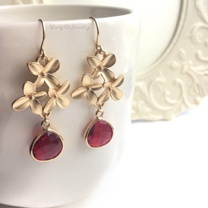 May include: Gold flower dangle earrings with red gemstone drops. The earrings feature a cluster of five gold flowers with a small center bead on each flower.