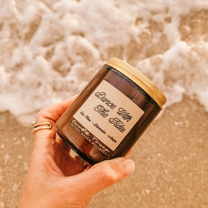 May include: A brown glass candle jar with a gold lid. The label reads "Dance With The Tides" and lists the scent as "Sea Salt - Lavender - Amber". The candle is labeled "Earth Transit" and "Nature Based Wellness".