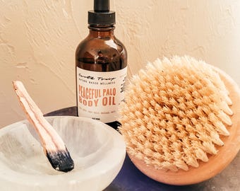 May include: A brown glass bottle with a dropper top, labeled "Peaceful Palo Body Oil" by Earth Trust. The bottle is next to a wooden brush with natural bristles and a white selenite bowl with a wooden stick inside.