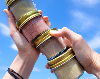 May include: Four glass jars with gold lids stacked on top of each other. The jars are filled with different coloured powders. The jars are being held by a person's hands against a blue sky background.