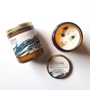 May include: Two amber glass candle jars with gold lids. The larger jar has a blue and white watercolor design of waves. The smaller jar has a label that reads "Design By JESSANNE Carth Tray FIR NEEDLE + LAVENDER + GUAIAC WOOD 50 HOUR BURN TIME 100% NATURAL SOY WAX INFUSED WITH ESSENTIAL OILS CRYSTAL & BOTANICALS".