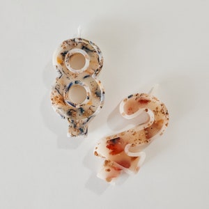 May include: Two white birthday candles, one shaped like the number 8 and the other shaped like the number 2. Both candles are decorated with a mix of gold, brown, and blue glitter.