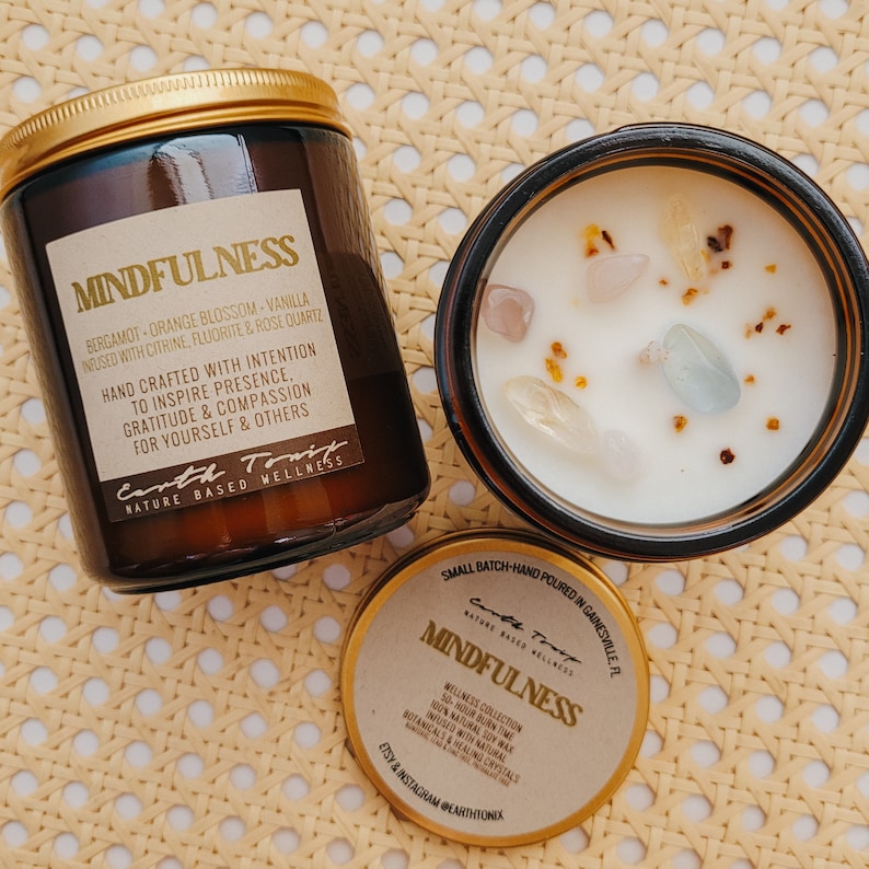 May include: A brown glass candle jar with a gold lid and label that reads "MINDFULNESS" with the text "BERGAMOT - ORANGE BLOSSOM - VANILLA INFUSED WITH CITRINE, FLUORITE & ROSE QUARTZ" and "HAND CRAFTED WITH INTENTION TO INSPIRE PRESENCE, GRATITUDE & COMPASSION FOR YOURSELF & OTHERS" and "Earth Tonic NATURE BASED WELLNESS". The candle jar is sitting on a woven straw surface. A smaller gold jar with a label that reads "MINDFULNESS" and "WELLNESS COLLECTION 50-HOUR BURN TIME 100% NATURAL SOY WAX INFUSED WITH NATURAL BOTANICALS & HEALING CRYSTALS" and "ETSY & INSTAGRAM @EARTHTONIK" is also visible.