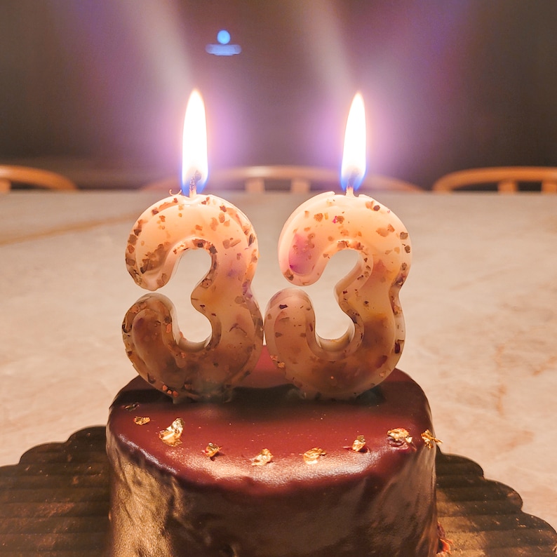 May include: Two lit candles shaped like the numbers 3 and 3 on top of a chocolate cake with gold leaf decorations.